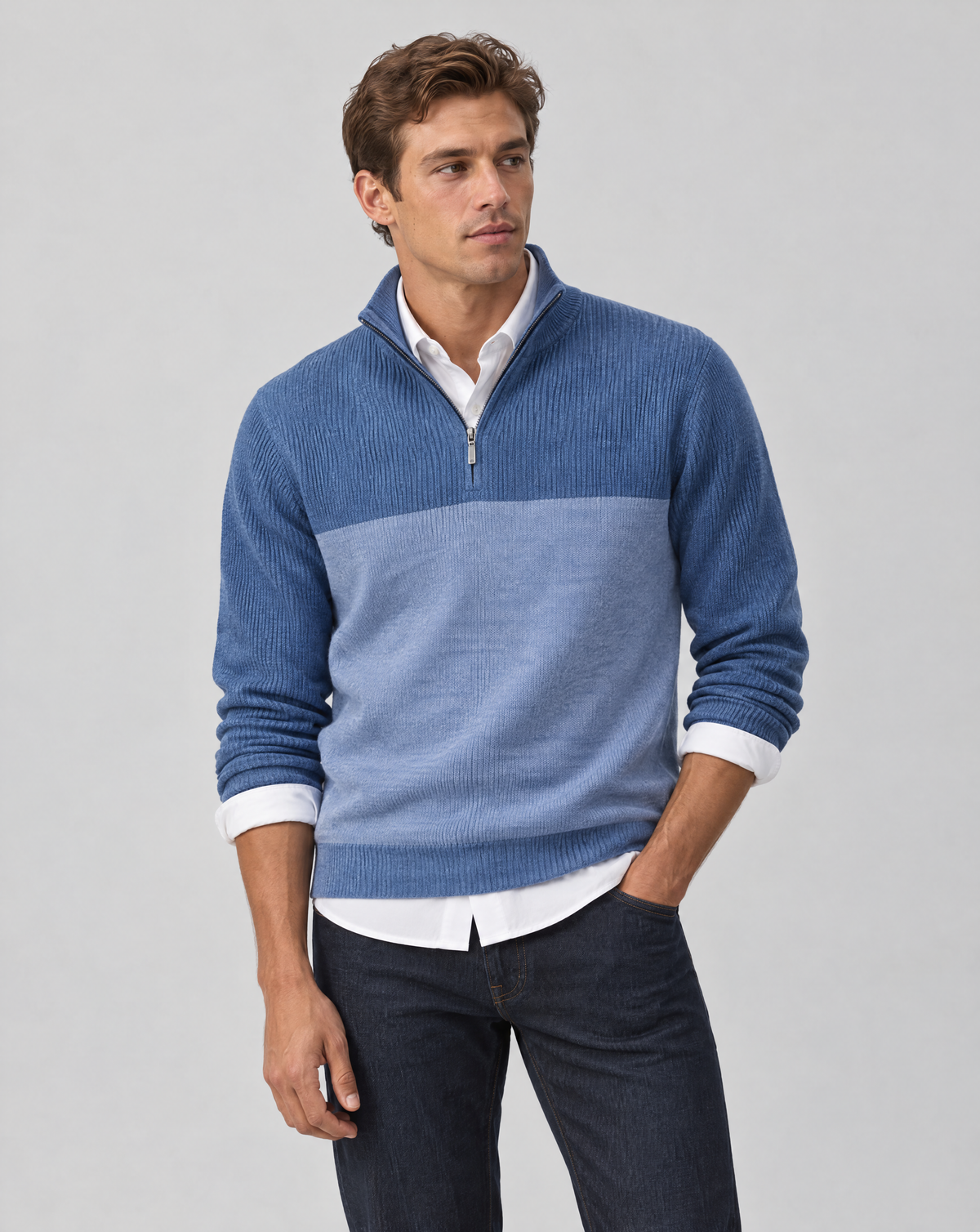 A man wearing a blue and light blue color-blocked quarter-zip sweater over a white collared shirt and dark jeans, standing against a light gray studio background.